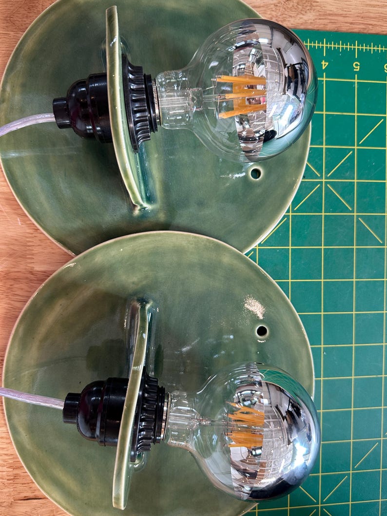 May include: Two wall sconces featuring circular, moss-green glazed ceramic backplates with a single mounting hole. Each fixture includes a black socket holding a large, clear globe-shaped LED filament light bulb with a silver-tinted top section.