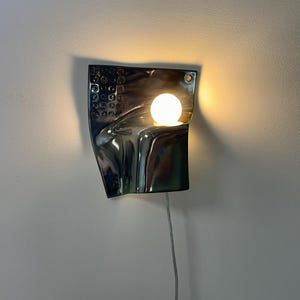 K&ouml;nnte beinhalten: Eine Wandleuchte mit einer reflektierenden, dunkelmetallischen, geschwungenen rechteckigen Platte, die auf der linken Seite ein strukturiertes Gittermuster aufweist. Eine einzelne, leuchtende kugelf&ouml;rmige Gl&uuml;hbirne wirft warmes Licht.
