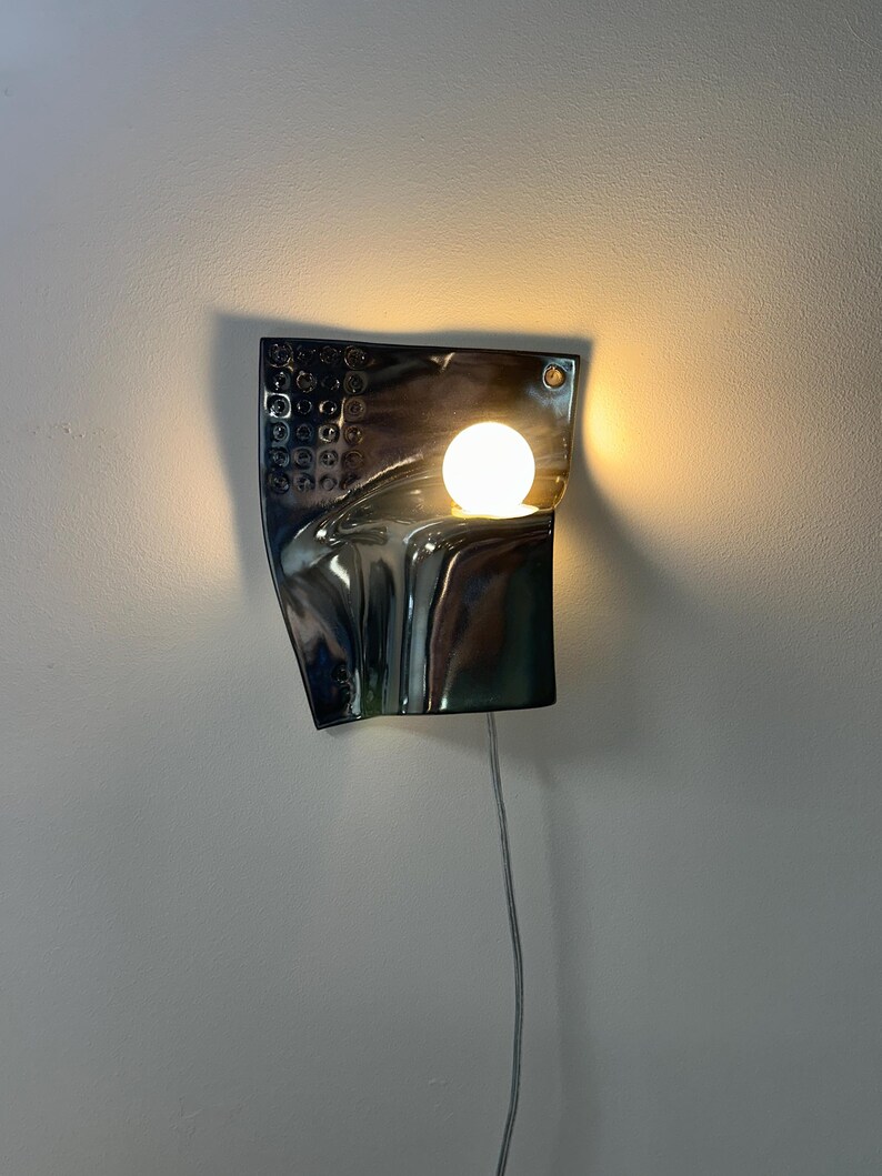 K&ouml;nnte beinhalten: Eine Wandleuchte mit einer reflektierenden, dunkelmetallischen, geschwungenen rechteckigen Platte, die auf der linken Seite ein strukturiertes Gittermuster aufweist. Eine einzelne, leuchtende kugelf&ouml;rmige Gl&uuml;hbirne wirft warmes Licht.