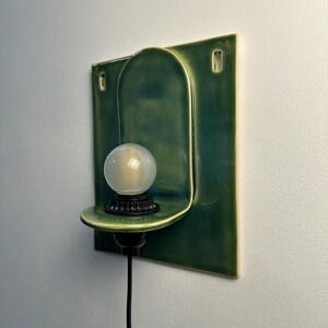 May include: A wall-mounted ceramic light fixture featuring a rectangular green backplate with an arched center section. A small, circular shelf extends from the center, holding a black socket and a round, frosted white glass light bulb with a black cord.