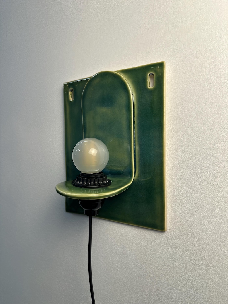 May include: A wall-mounted ceramic light fixture featuring a rectangular green backplate with an arched center section. A small, circular shelf extends from the center, holding a black socket and a round, frosted white glass light bulb with a black cord.
