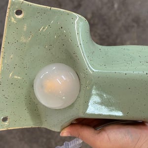 May include: A sage green ceramic wall sconce with a speckled finish and a glossy glaze. The fixture features a flat mounting plate with two circular holes for installation and a protruding cylindrical section that houses a round, frosted white light bulb.