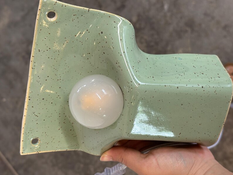 May include: A sage green ceramic wall sconce with a speckled finish and a glossy glaze. The fixture features a flat mounting plate with two circular holes for installation and a protruding cylindrical section that houses a round, frosted white light bulb.