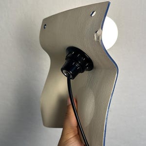 May include: A wall-mounted light fixture featuring a curved, beige textured panel with two small circular holes at the top. A black socket is centered on the panel, holding a white spherical light bulb, with a black power cord extending downward.