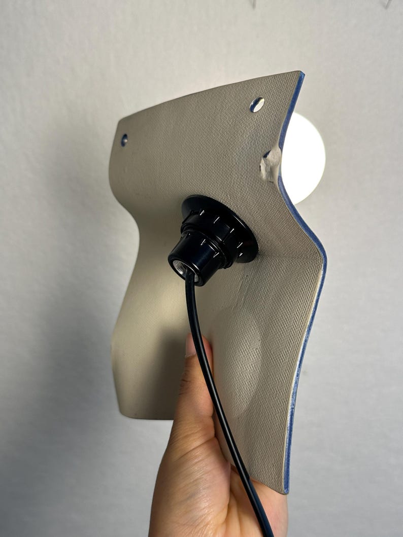 May include: A wall-mounted light fixture featuring a curved, beige textured panel with two small circular holes at the top. A black socket is centered on the panel, holding a white spherical light bulb, with a black power cord extending downward.