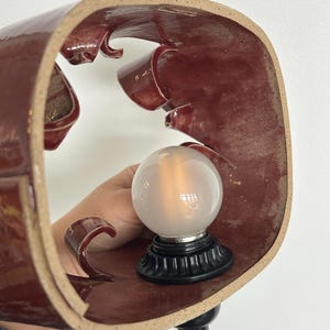 May include: A maroon ceramic lamp with a unique, curved design. The lamp features a frosted glass bulb and a black base. The lamp is held by a person's hand. The lamp is plugged into a black cord.