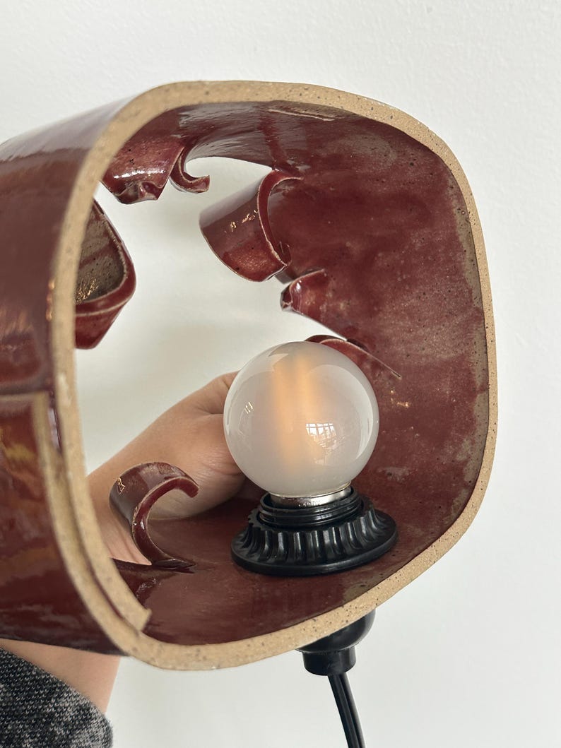 May include: A maroon ceramic lamp with a unique, curved design. The lamp features a frosted glass bulb and a black base. The lamp is held by a person's hand. The lamp is plugged into a black cord.