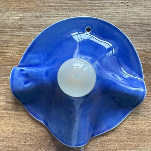 May include: A blue ceramic wall sconce with a white light bulb in the center. The sconce has a wavy, organic shape and a glossy finish. A small hole is at the top for hanging. A white cord extends from the bottom.