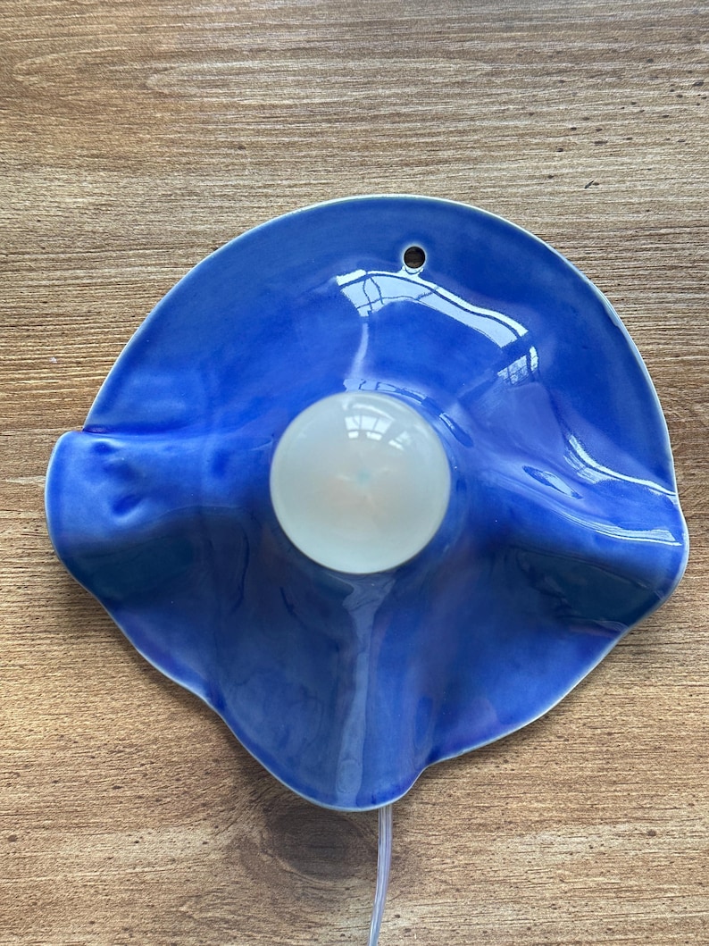 May include: A blue ceramic wall sconce with a white light bulb in the center. The sconce has a wavy, organic shape and a glossy finish. A small hole is at the top for hanging. A white cord extends from the bottom.