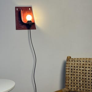 May include: A wall-mounted lamp featuring a rectangular, deep red ceramic backplate with a curved, shell-like form. A black socket holds a glowing, round light bulb, and a thin black power cord hangs vertically downward from the fixture.