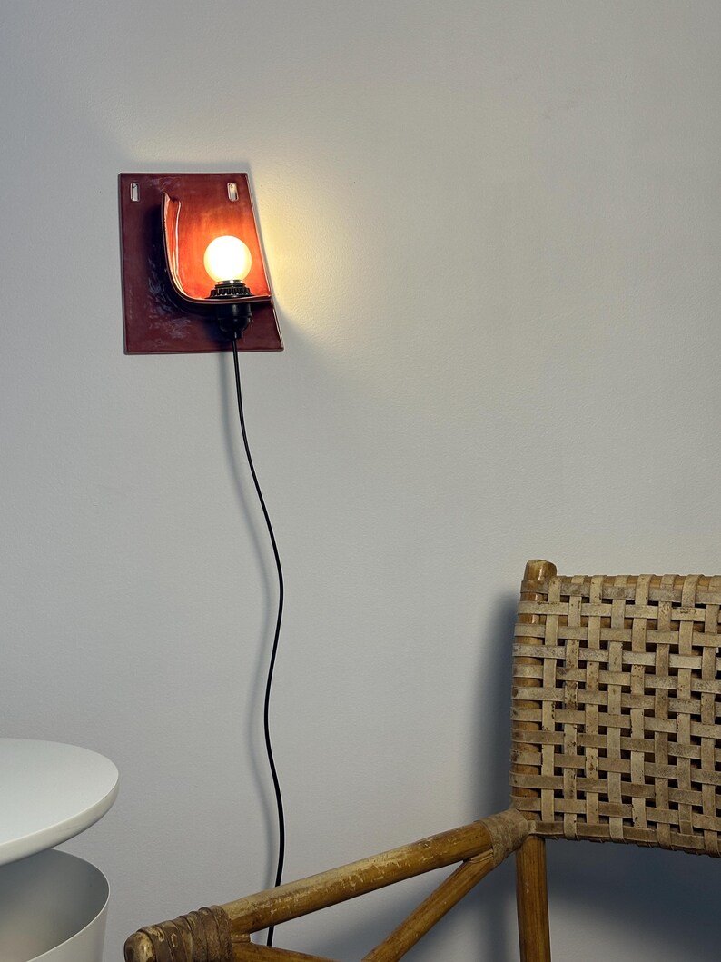 May include: A wall-mounted lamp featuring a rectangular, deep red ceramic backplate with a curved, shell-like form. A black socket holds a glowing, round light bulb, and a thin black power cord hangs vertically downward from the fixture.