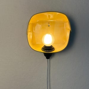 May include: A wall-mounted lamp featuring a bright yellow, square-shaped ceramic shade with rounded corners. A single glowing white light bulb is centered within the shade, attached to a black socket base with a thin white power cord hanging below.