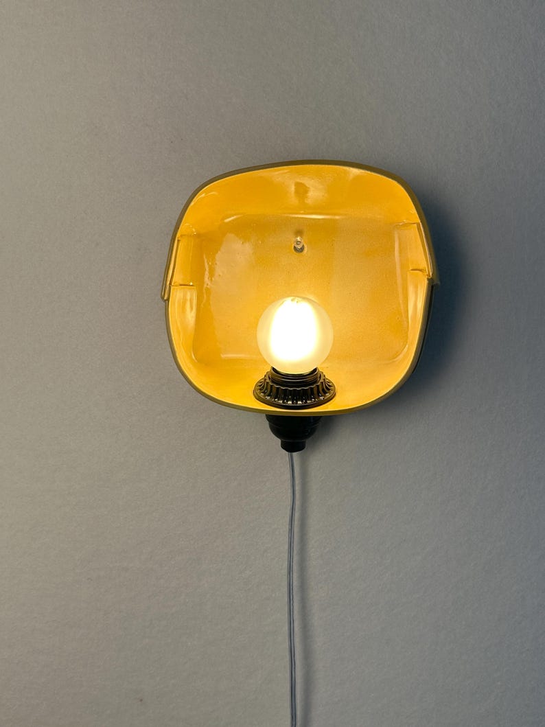 May include: A wall-mounted lamp featuring a bright yellow, square-shaped ceramic shade with rounded corners. A single glowing white light bulb is centered within the shade, attached to a black socket base with a thin white power cord hanging below.