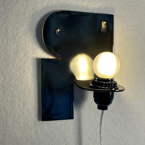 May include: A wall-mounted lamp featuring a glossy, dark navy blue ceramic backplate with an irregular, geometric shape. A black socket holds a single, glowing, frosted spherical light bulb, with a thin, clear power cord extending downwards.