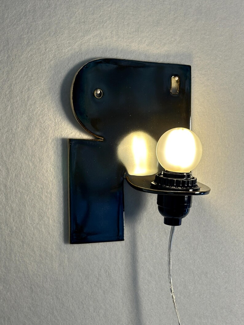 May include: A wall-mounted lamp featuring a glossy, dark navy blue ceramic backplate with an irregular, geometric shape. A black socket holds a single, glowing, frosted spherical light bulb, with a thin, clear power cord extending downwards.