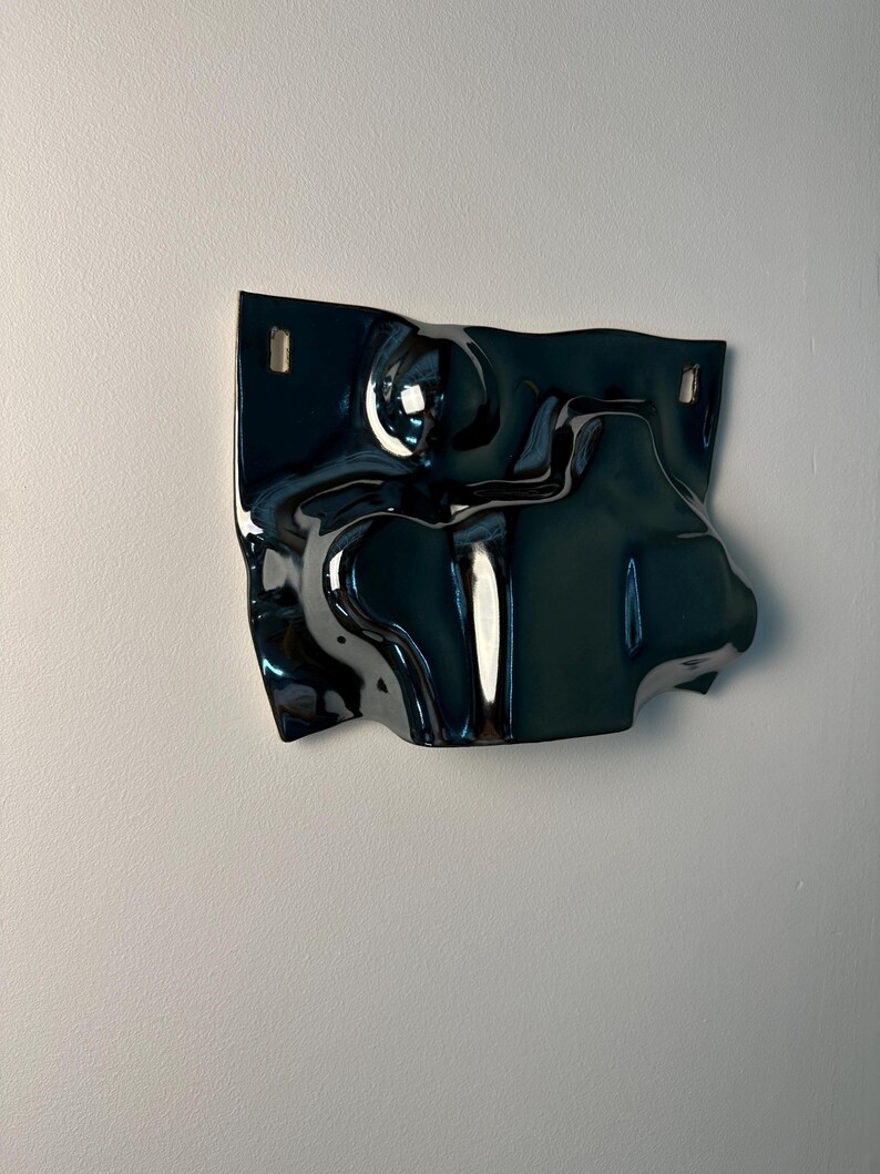 Pu&ograve; includere: Un oggetto scultoreo da parete, scuro e lucido, caratterizzato da una superficie fluida e ondulata. Il pezzo presenta una finitura profonda e riflettente e due aperture rettangolari vicino al bordo superiore per il fissaggio.
