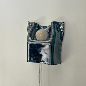 May include: A wall-mounted lamp featuring a reflective, chrome-finished ceramic base with an irregular, fluid shape. A single, round, frosted white light bulb is centered within the recessed upper section, and a thin, clear power cord hangs below.