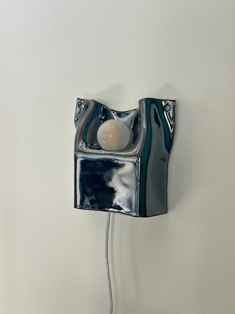May include: A wall-mounted lamp featuring a reflective, chrome-finished ceramic base with an irregular, fluid shape. A single, round, frosted white light bulb is centered within the recessed upper section, and a thin, clear power cord hangs below.