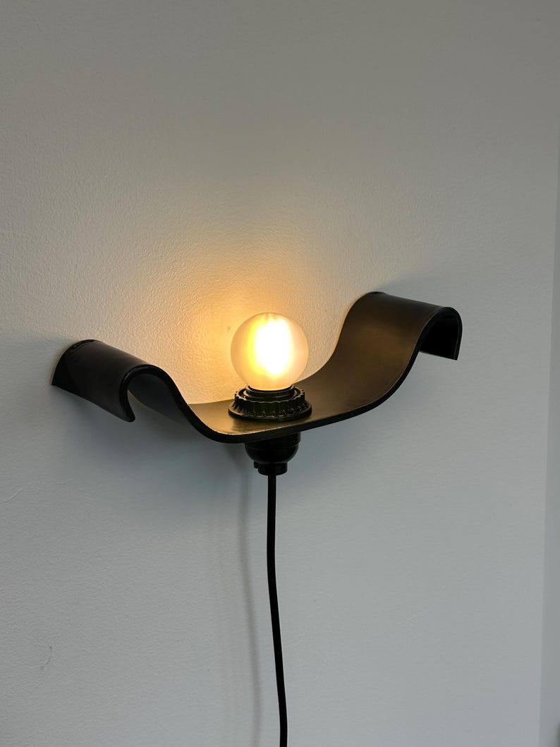 May include: A black wall-mounted light fixture with a curved, wave-like design. The fixture holds a round, glowing light bulb. A black cord extends downward from the fixture.