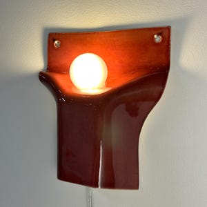 Pode incluir: Um candeeiro de parede em cer&acirc;mica com um acabamento vermelho escuro brilhante e uma forma escultural curva. A lumin&aacute;ria inclui uma abertura circular central com uma l&acirc;mpada esf&eacute;rica branca brilhante e dois orif&iacute;cios superiores para fixa&ccedil;&atilde;o.