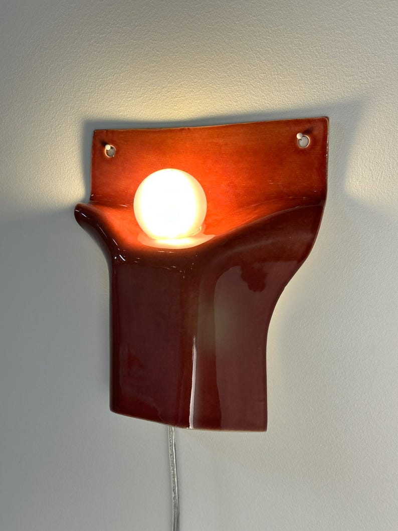 Pode incluir: Um candeeiro de parede em cer&acirc;mica com um acabamento vermelho escuro brilhante e uma forma escultural curva. A lumin&aacute;ria inclui uma abertura circular central com uma l&acirc;mpada esf&eacute;rica branca brilhante e dois orif&iacute;cios superiores para fixa&ccedil;&atilde;o.