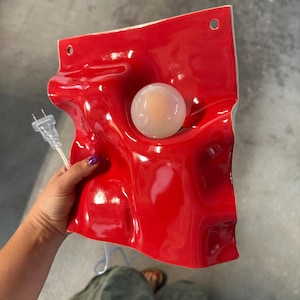 May include: A glossy red ceramic wall lamp with an undulating, wave-like surface. The fixture features a central circular opening holding a round, frosted white light bulb. Two small holes are positioned at the top for mounting, and a clear electrical cord with a standard two-prong plug extends from the side.