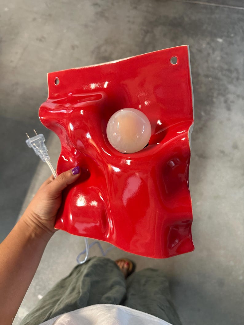 May include: A glossy red ceramic wall lamp with an undulating, wave-like surface. The fixture features a central circular opening holding a round, frosted white light bulb. Two small holes are positioned at the top for mounting, and a clear electrical cord with a standard two-prong plug extends from the side.