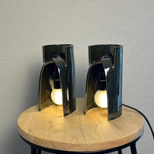 Puede incluir: Dos l&aacute;mparas de mesa de cer&aacute;mica verde oscuro con una silueta curva y arquitect&oacute;nica. Cada l&aacute;mpara presenta un acabado brillante y una estructura abierta que alberga una bombilla blanca brillante suspendida de un casquillo negro.
