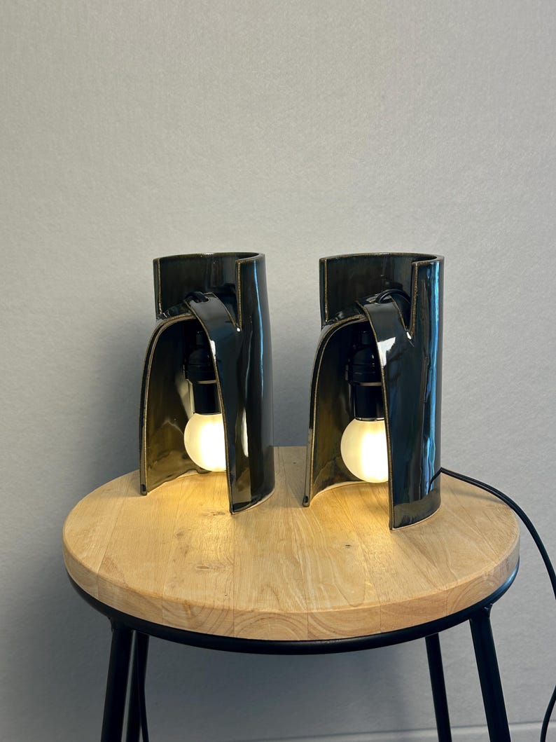 Puede incluir: Dos l&aacute;mparas de mesa de cer&aacute;mica verde oscuro con una silueta curva y arquitect&oacute;nica. Cada l&aacute;mpara presenta un acabado brillante y una estructura abierta que alberga una bombilla blanca brillante suspendida de un casquillo negro.