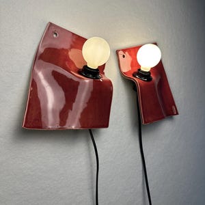 Puede incluir: Dos l&aacute;mparas de pared con pantallas de cer&aacute;mica de color rojo oscuro brillante y bordes irregulares y curvos. Cada l&aacute;mpara tiene una bombilla blanca redonda y brillante en un casquillo negro, con un cable el&eacute;ctrico negro hacia abajo.