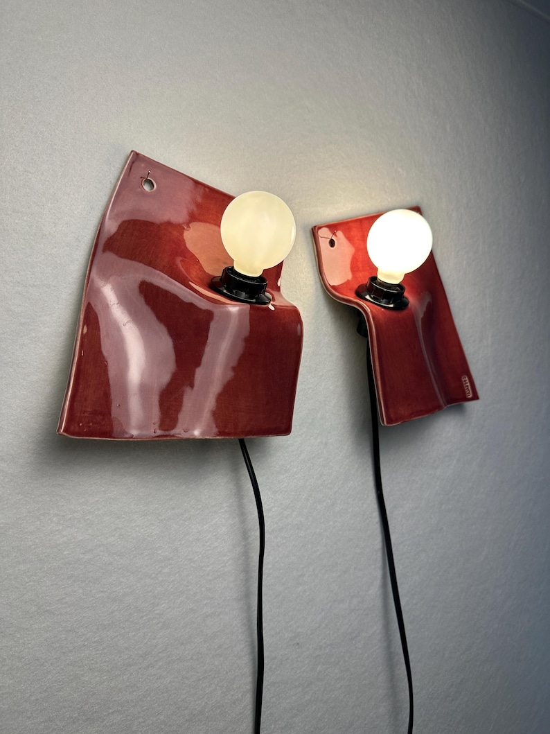 Puede incluir: Dos l&aacute;mparas de pared con pantallas de cer&aacute;mica de color rojo oscuro brillante y bordes irregulares y curvos. Cada l&aacute;mpara tiene una bombilla blanca redonda y brillante en un casquillo negro, con un cable el&eacute;ctrico negro hacia abajo.