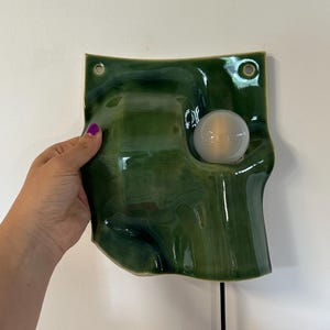 May include: A wall-mounted lamp featuring a glossy, dark green ceramic shade with an irregular, wavy surface. The shade has two circular holes at the top corners for mounting and a single round, white light bulb protruding from the right side.
