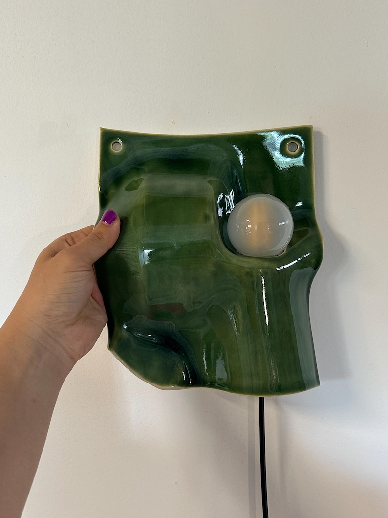May include: A wall-mounted lamp featuring a glossy, dark green ceramic shade with an irregular, wavy surface. The shade has two circular holes at the top corners for mounting and a single round, white light bulb protruding from the right side.