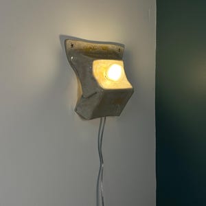 May include: A wall-mounted ceramic light fixture with a glossy off-white finish and a sculptural, folded form. The fixture features a single glowing light bulb centered in a recessed area, with a clear electrical cord extending downward from the base.