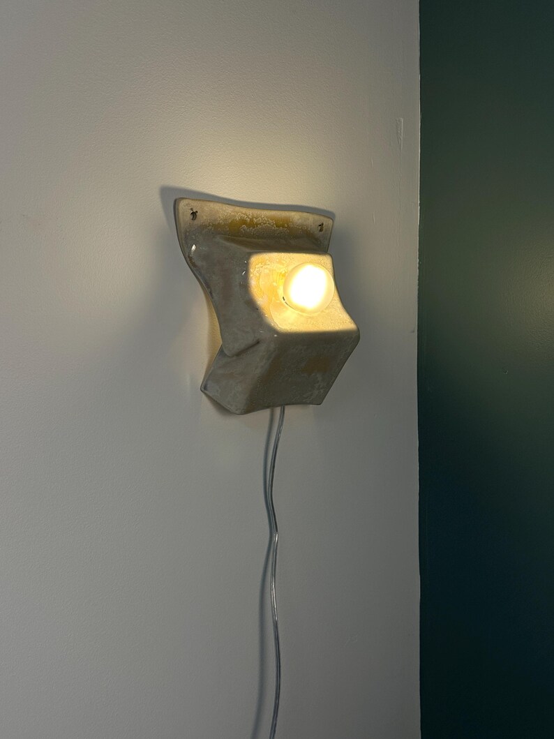 May include: A wall-mounted ceramic light fixture with a glossy off-white finish and a sculptural, folded form. The fixture features a single glowing light bulb centered in a recessed area, with a clear electrical cord extending downward from the base.