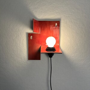 Pode incluir: Uma arandela de parede vermelha moderna com um design geom&eacute;trico &uacute;nico. A arandela apresenta uma pequena prateleira que suporta uma l&acirc;mpada redonda e branca. Um cabo preto estende-se para baixo. A arandela &eacute; montada numa parede cinzenta.