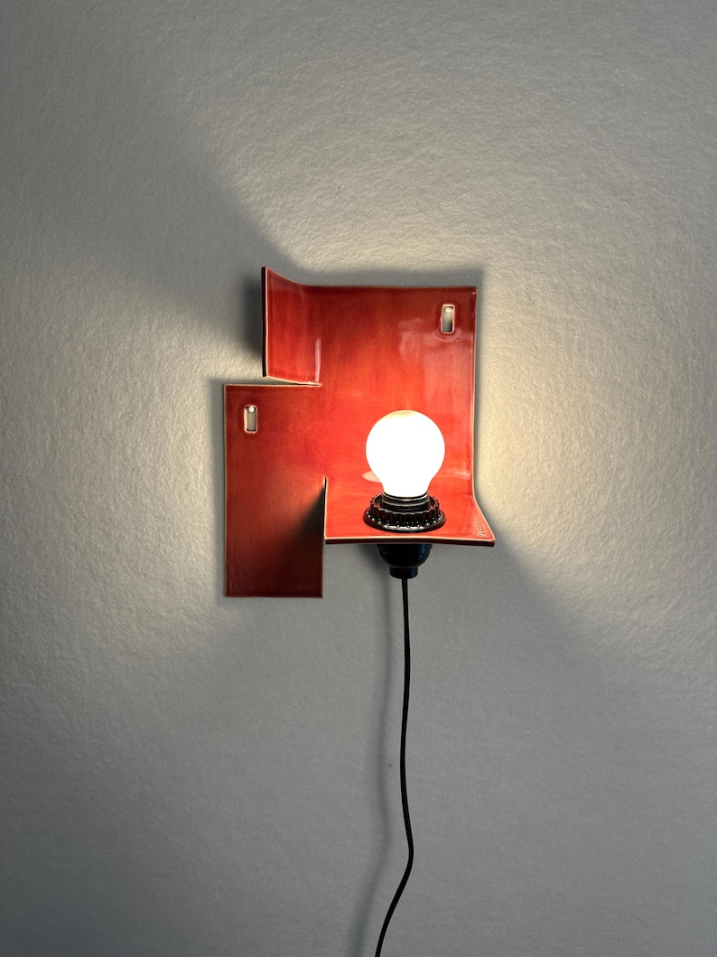 Pode incluir: Uma arandela de parede vermelha moderna com um design geom&eacute;trico &uacute;nico. A arandela apresenta uma pequena prateleira que suporta uma l&acirc;mpada redonda e branca. Um cabo preto estende-se para baixo. A arandela &eacute; montada numa parede cinzenta.