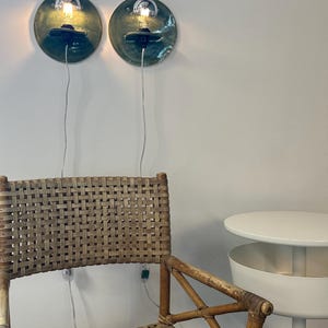 May include: Two circular wall-mounted lamps with dark green glazed ceramic backings and exposed light bulbs. Below them sits a woven rattan chair with a high back and a white side table with a cylindrical base and a circular top surface.