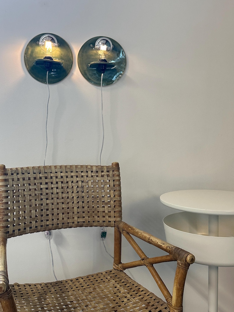May include: Two circular wall-mounted lamps with dark green glazed ceramic backings and exposed light bulbs. Below them sits a woven rattan chair with a high back and a white side table with a cylindrical base and a circular top surface.