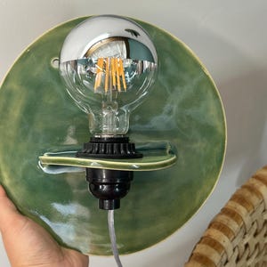 May include: A circular wall sconce featuring a glossy sage green ceramic backplate with a small integrated shelf. A black socket holds a clear glass light bulb with a reflective silver top half and visible internal vertical filaments.