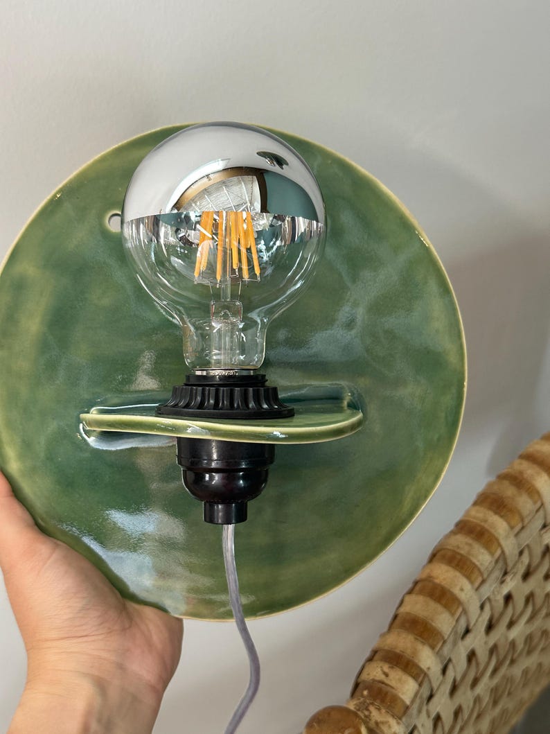 May include: A circular wall sconce featuring a glossy sage green ceramic backplate with a small integrated shelf. A black socket holds a clear glass light bulb with a reflective silver top half and visible internal vertical filaments.