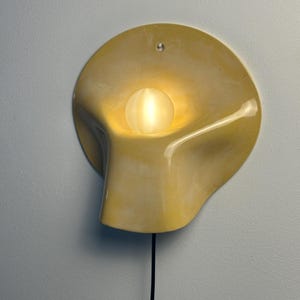 May include: A wall-mounted light fixture featuring a smooth, pale yellow, organic-shaped ceramic shade with a central circular opening. A small, frosted white spherical light bulb sits within the center, emitting a warm glow. A black power cord hangs down.