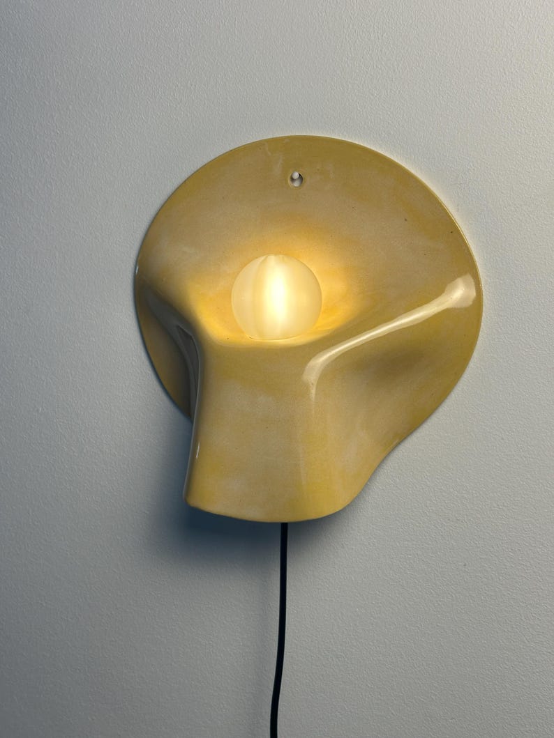 May include: A wall-mounted light fixture featuring a smooth, pale yellow, organic-shaped ceramic shade with a central circular opening. A small, frosted white spherical light bulb sits within the center, emitting a warm glow. A black power cord hangs down.