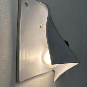 May include: A white ceramic wall sconce with a curved, sculptural form that extends outward. The fixture features a single glowing light bulb nestled within the concave shape, casting a soft light against the wall surface behind the fixture.
