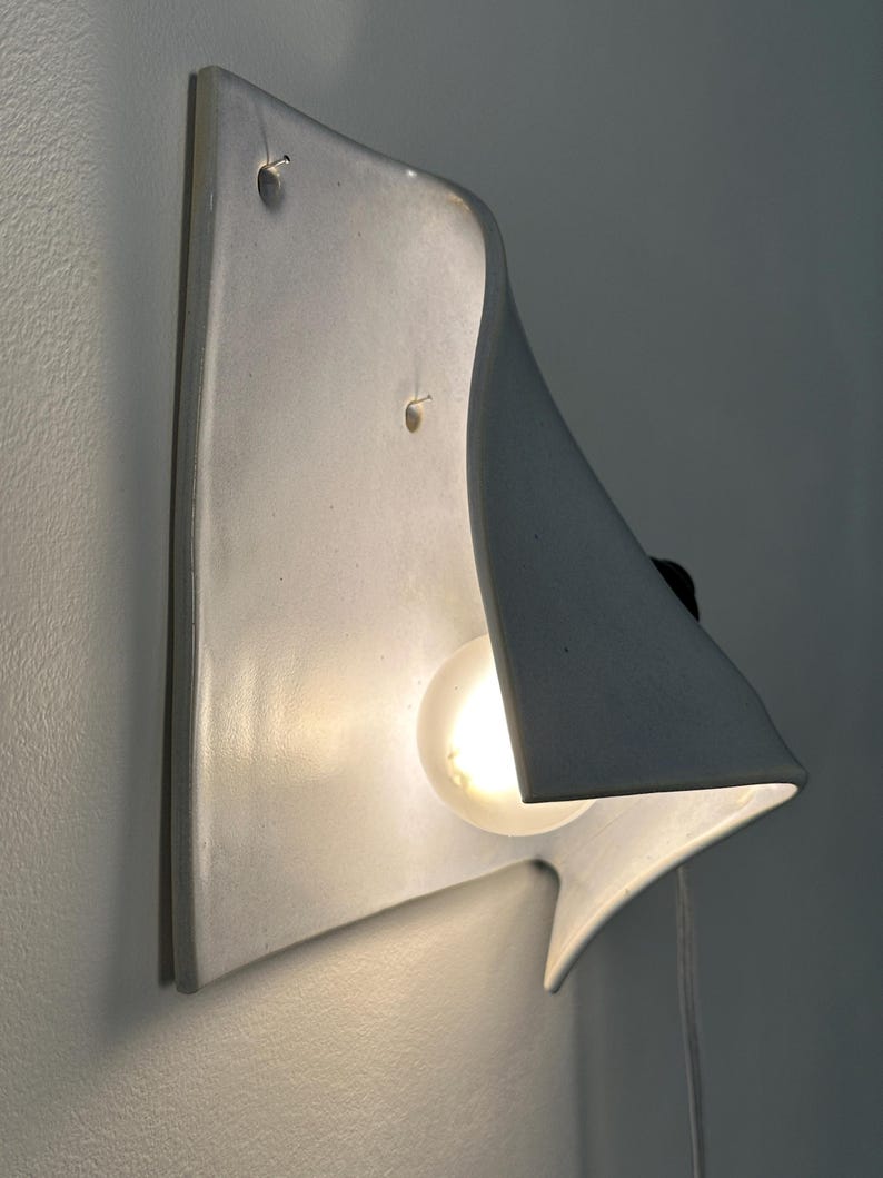 May include: A white ceramic wall sconce with a curved, sculptural form that extends outward. The fixture features a single glowing light bulb nestled within the concave shape, casting a soft light against the wall surface behind the fixture.