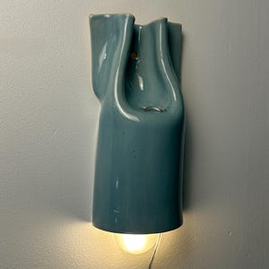 May include: A wall-mounted light fixture with a glossy, light blue ceramic shade featuring a fluid, organic, and sculptural form. The fixture emits a warm, downward-facing glow from a bulb positioned at the base, with a visible power cord extending.