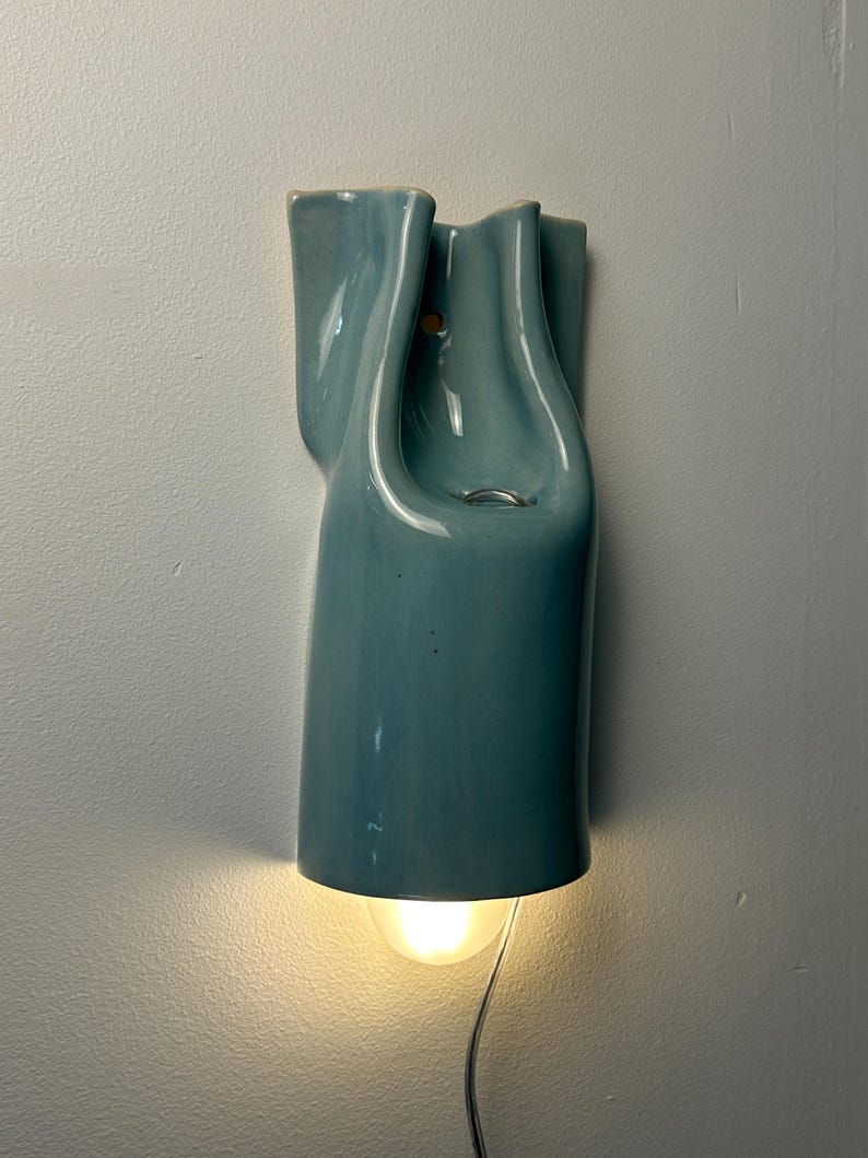 May include: A wall-mounted light fixture with a glossy, light blue ceramic shade featuring a fluid, organic, and sculptural form. The fixture emits a warm, downward-facing glow from a bulb positioned at the base, with a visible power cord extending.