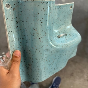 May include: A light blue ceramic wall sconce with a speckled brown pattern. The fixture features a curved, asymmetrical shape with two circular mounting holes at the top corners and a small metal loop on the front. A clear electrical plug is attached.