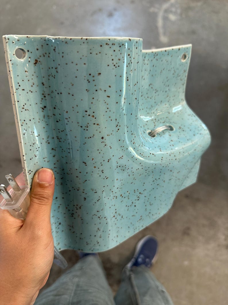 May include: A light blue ceramic wall sconce with a speckled brown pattern. The fixture features a curved, asymmetrical shape with two circular mounting holes at the top corners and a small metal loop on the front. A clear electrical plug is attached.
