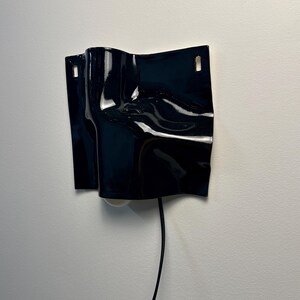 Puede incluir: Un aplique de pared negro brillante con una superficie curva y ondulada, que presenta dos ranuras de montaje rectangulares en la parte superior. La l&aacute;mpara tiene una bombilla visible en la parte inferior y un cable negro hacia abajo.